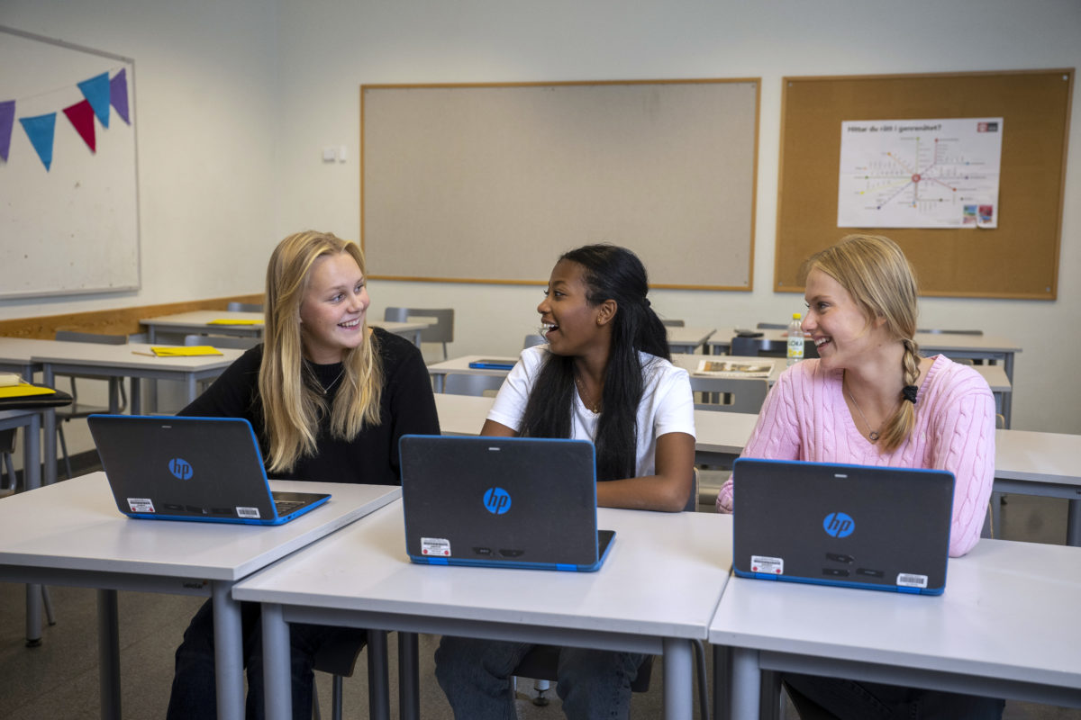 Tre glada tjejer diskuterar i skolbänken med en varsin laptop framför sig.